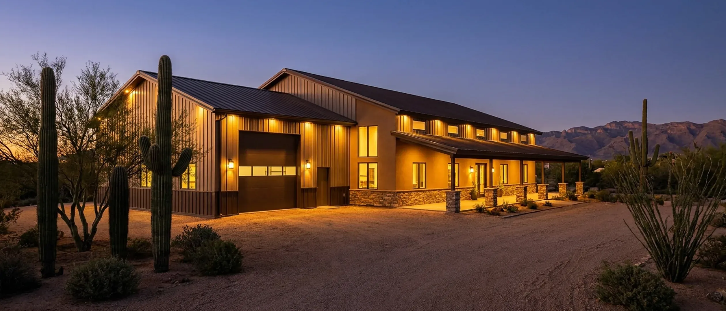 A barndominium at dusk on desert acreage, warm light in every window, with saguaro silhouettes and a mountain range on the horizon.